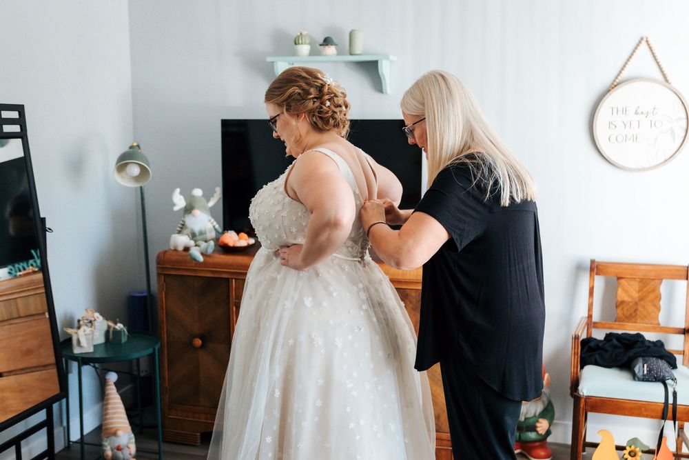 A woman in a lace wedding gown gets her dress fastened by another woman in a cozy home setting.