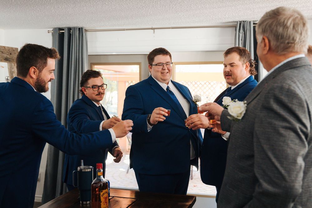 Groom and groomsmen in navy suits laughing and celebrating together before a wedding ceremony indoors.