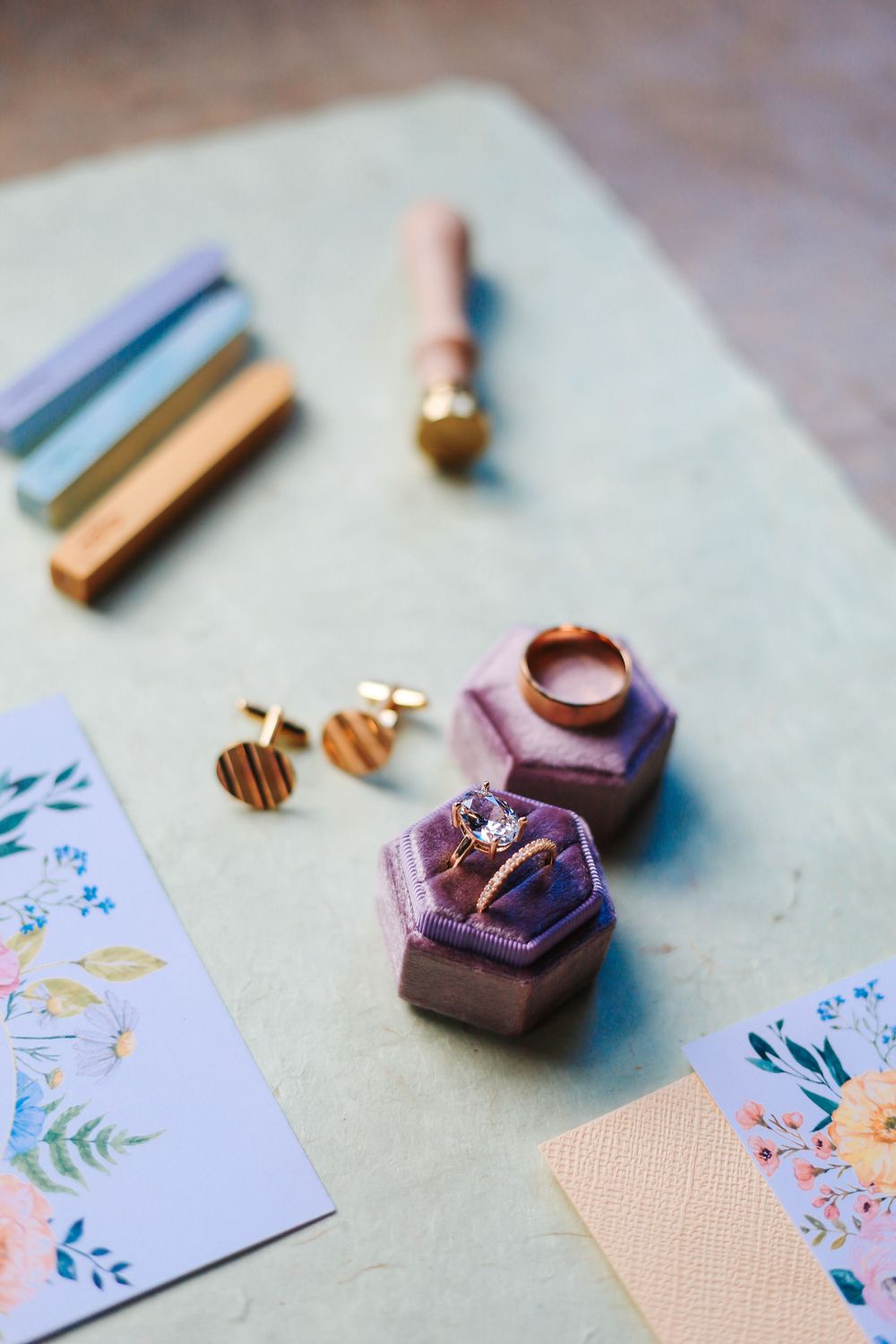 Gold engagement ring and band in purple velvet box with cufflinks, wax seal stamp, and floral cards on blue surface.