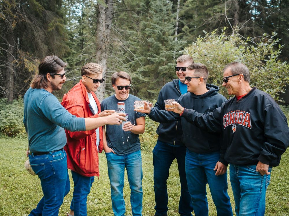 Six men toasting drinks outdoors in a forest setting, wearing casual clothes and sunglasses, enjoying a social gathering.