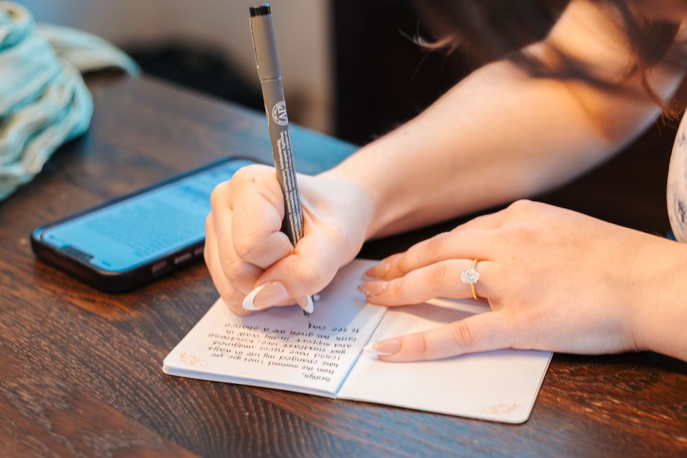 Woman writing in a small notebook with a pen, wearing a diamond ring, with a smartphone on a wooden table.