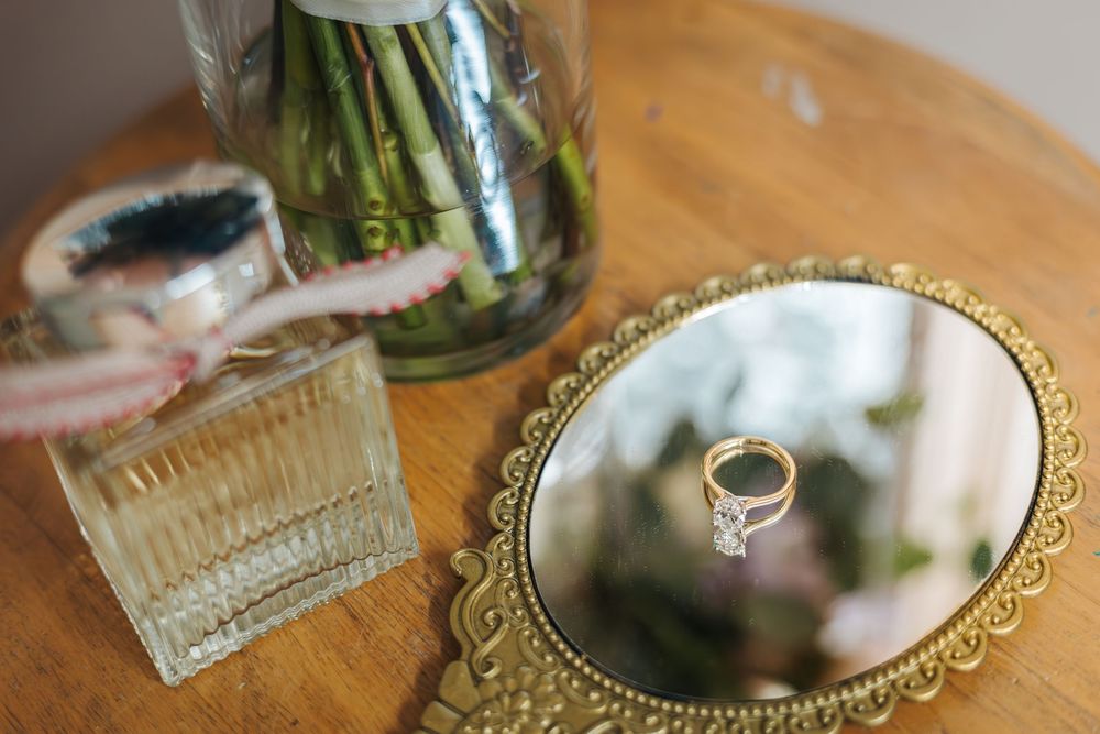 Gold oval mirror tray displaying a diamond engagement ring beside a perfume bottle and floral arrangement on a wooden table.