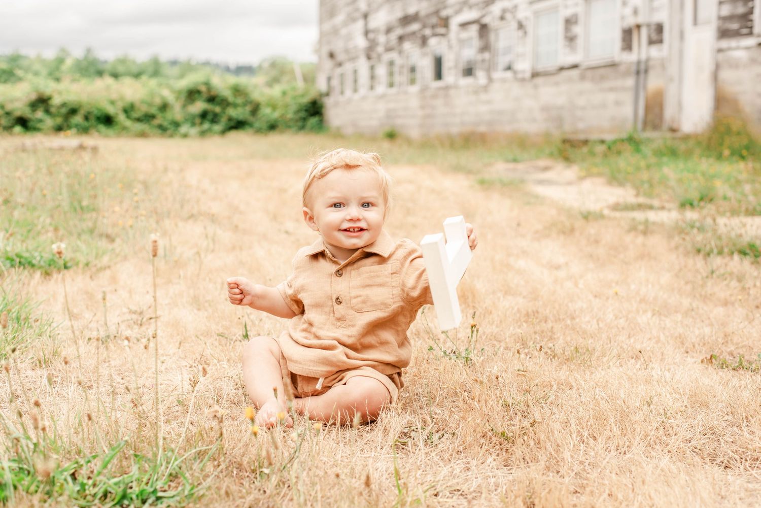 Happy baby in mustard romper sitting in golden grass field, smiling outdoors near rustic building.