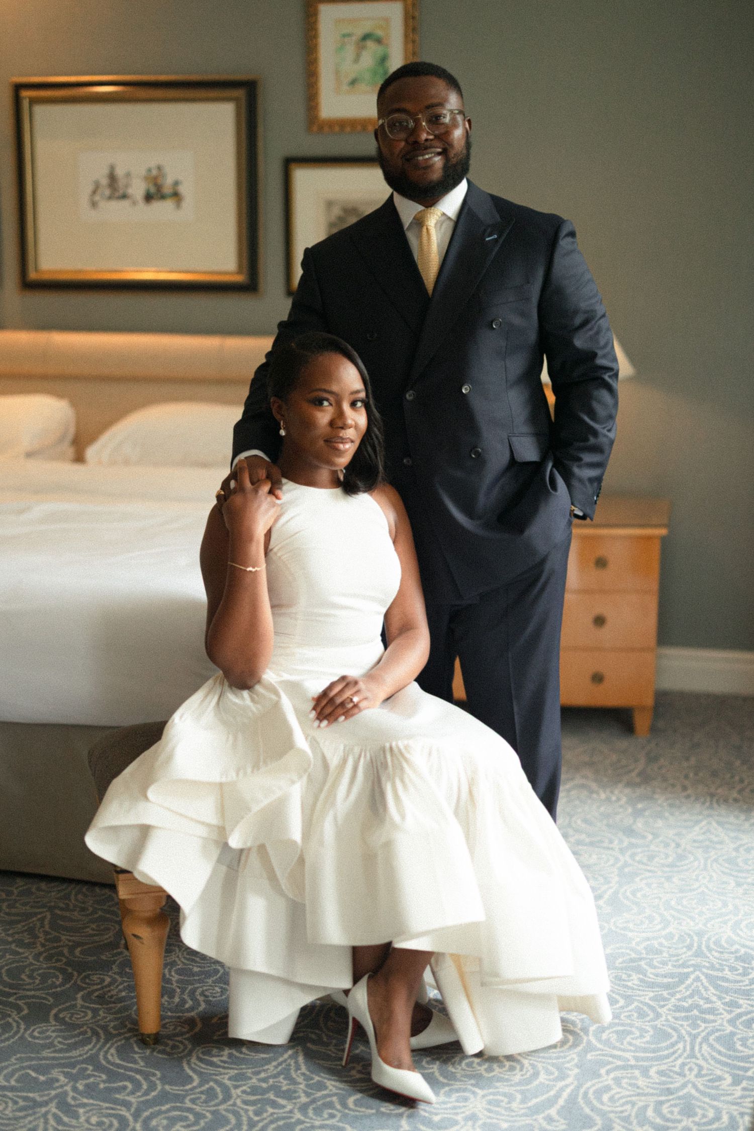 A couple in formal wedding attire poses together on a patterned carpet in a room with framed artwork.