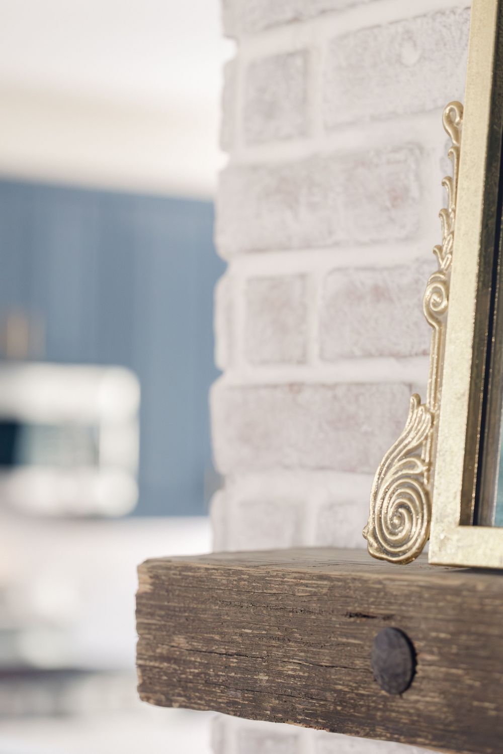 Close-up of a rustic wood fireplace mantel with ornate gold frame against a whitewashed brick surround.