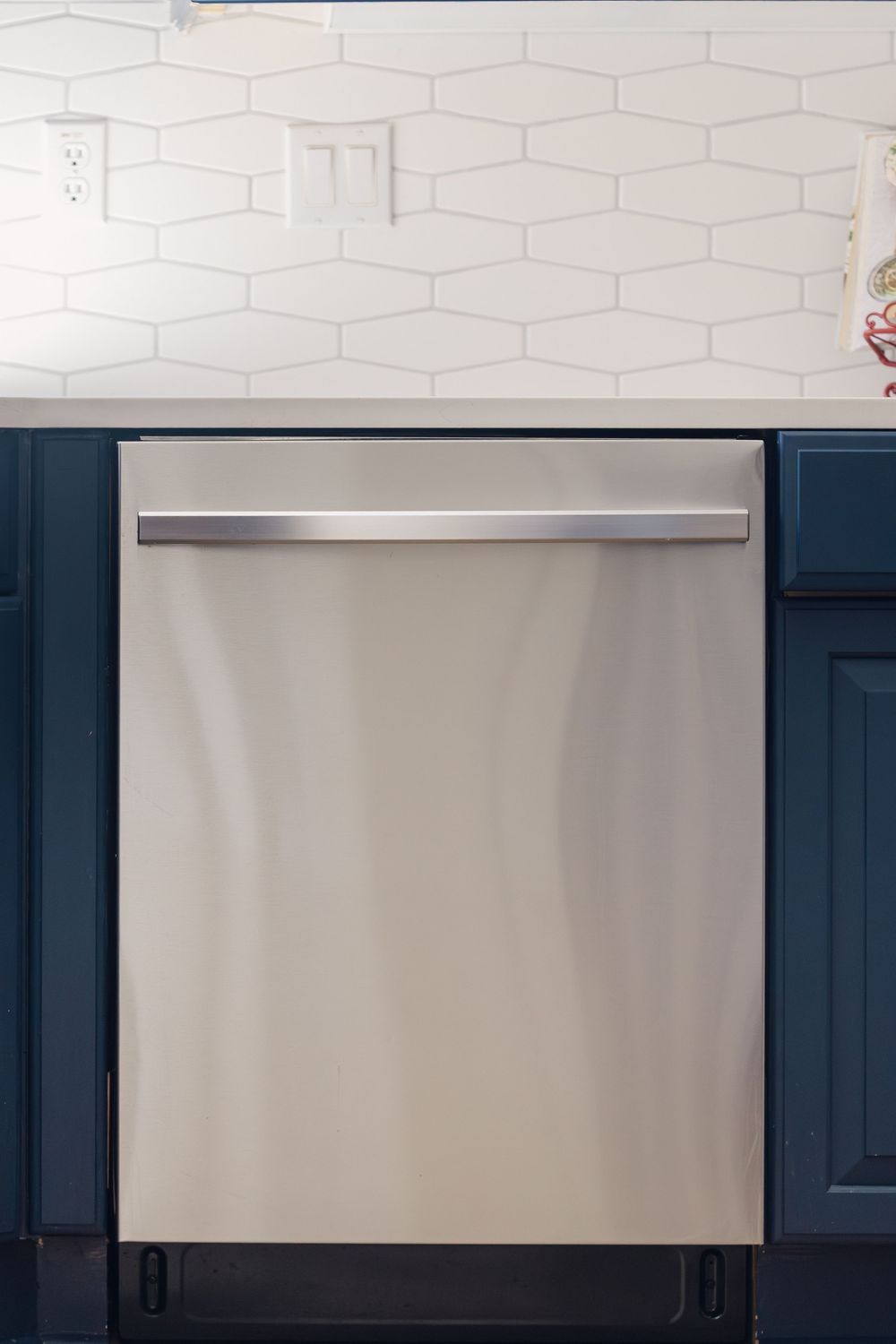 Stainless steel built-in dishwasher with navy blue cabinetry and white hexagonal tile backsplash in a modern kitchen.