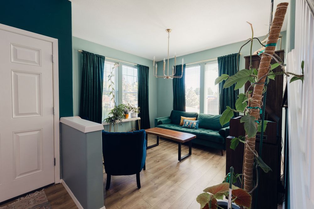 Modern living room with teal walls, green velvet sofa, wooden coffee table, and large window.