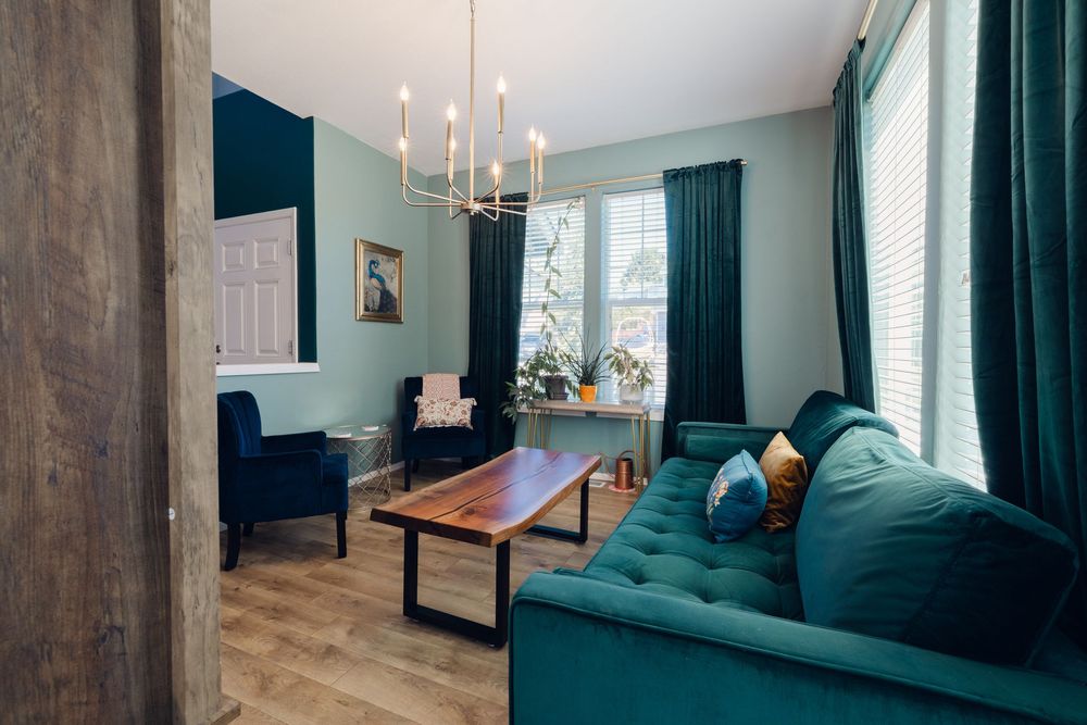 Elegant living room with teal velvet sofa, wooden coffee table, chandelier, and dark green curtains on bright windows.