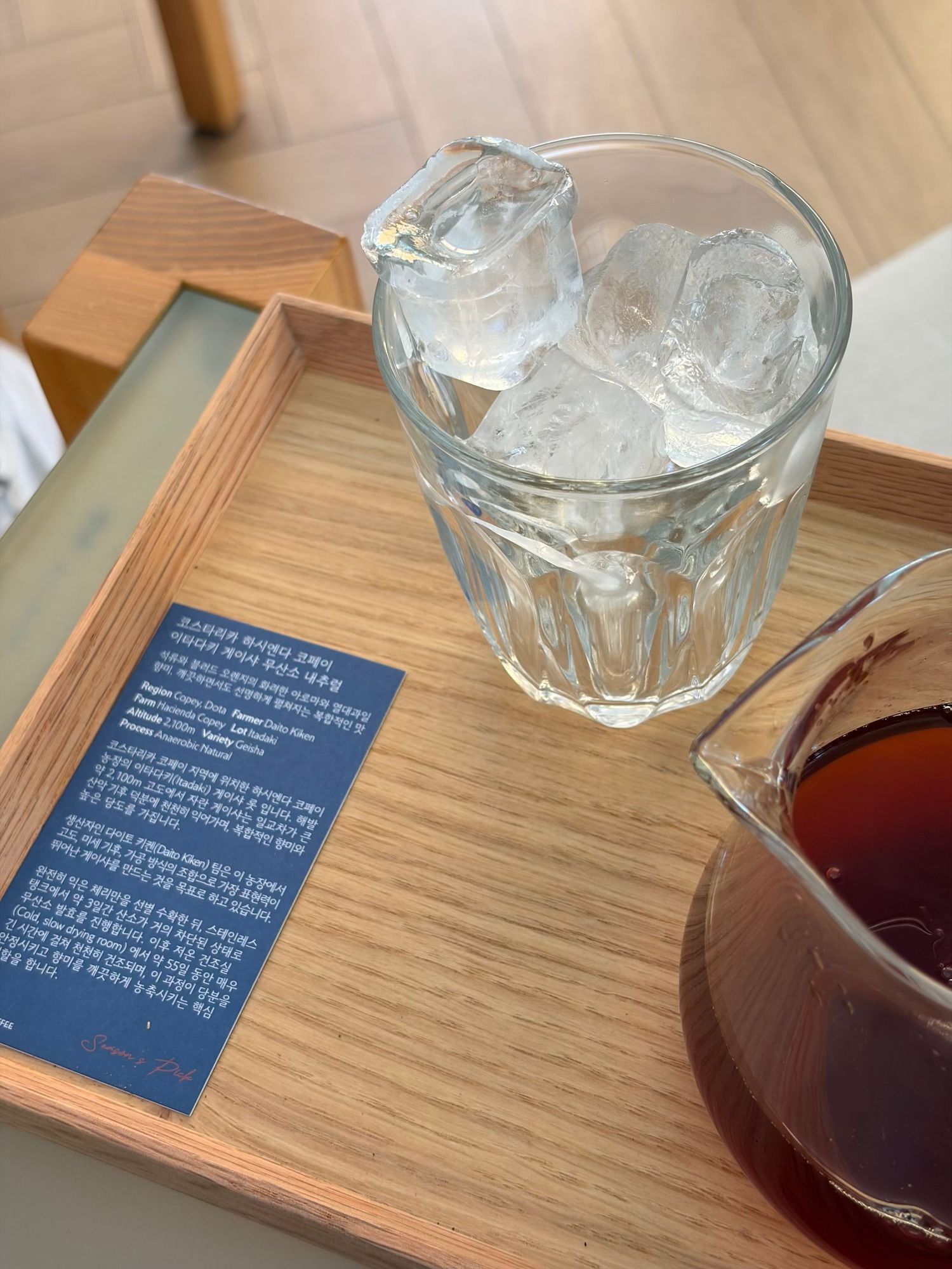 Wooden tray with glass of ice, Korean menu card, and pitcher of black coffee on cafe table.