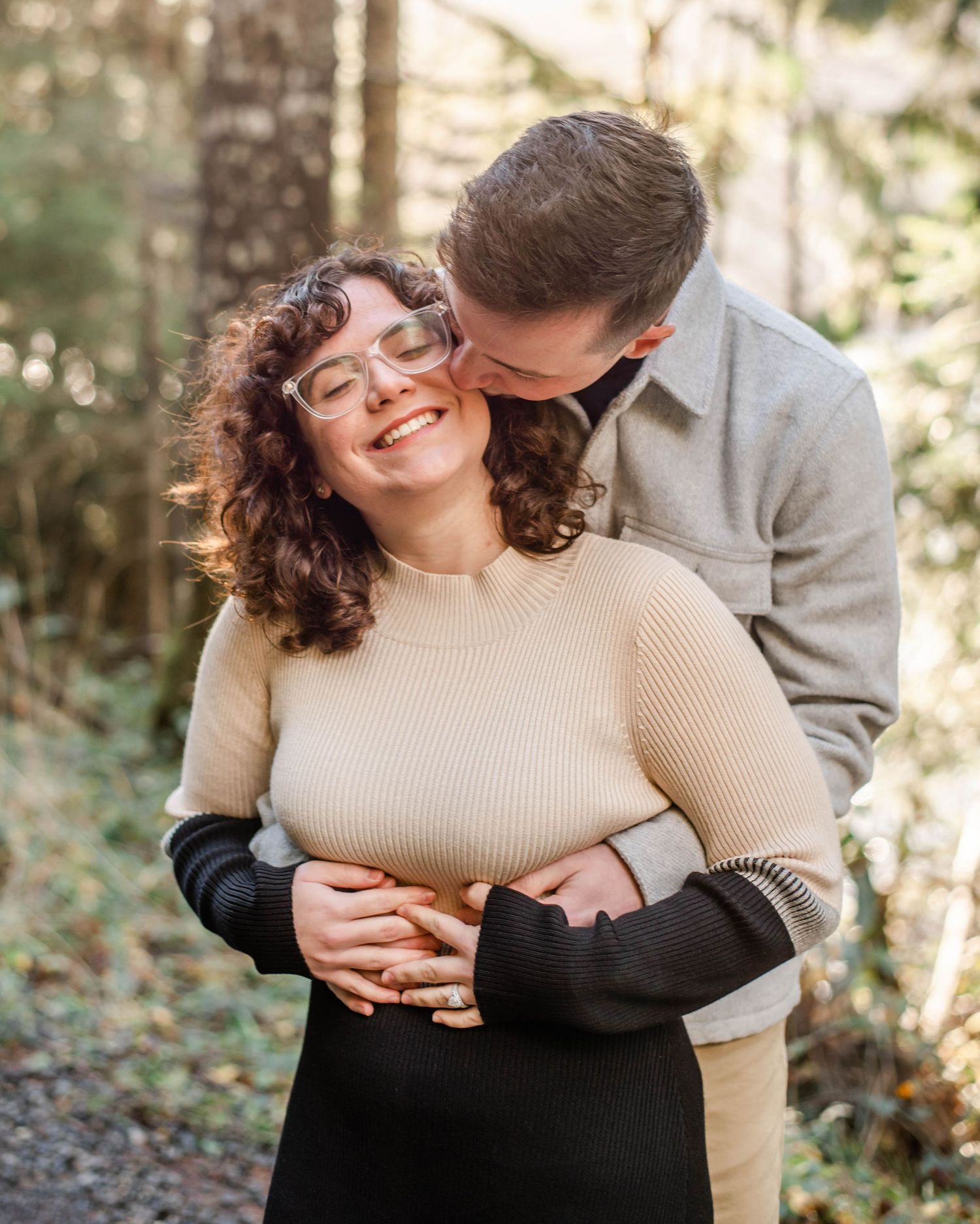 A couple embraces outdoors in a forest, woman smiling joyfully as man kisses her temple in a tender moment.