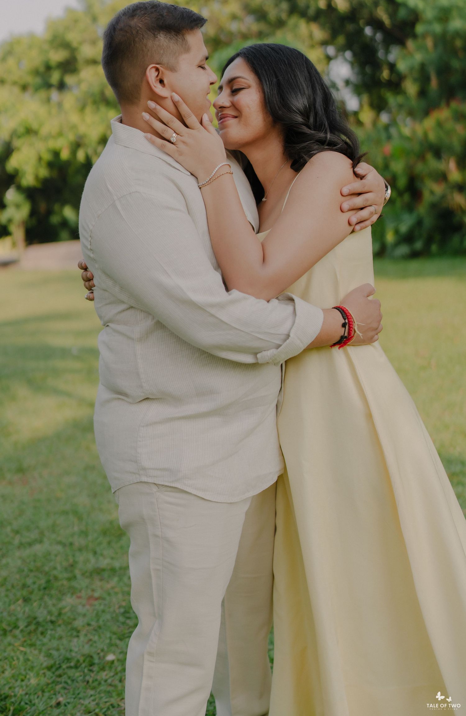 A couple in matching cream outfits embrace and smile at each other in a lush green park setting.