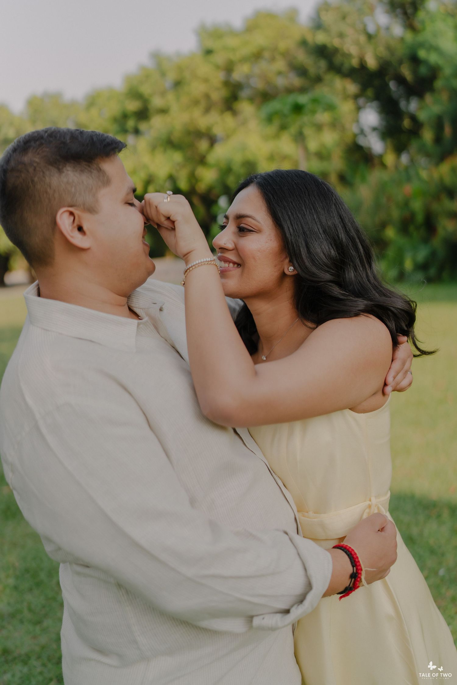 A couple shares a romantic moment outdoors, with the woman smiling as the man kisses her hand in a lush green park.