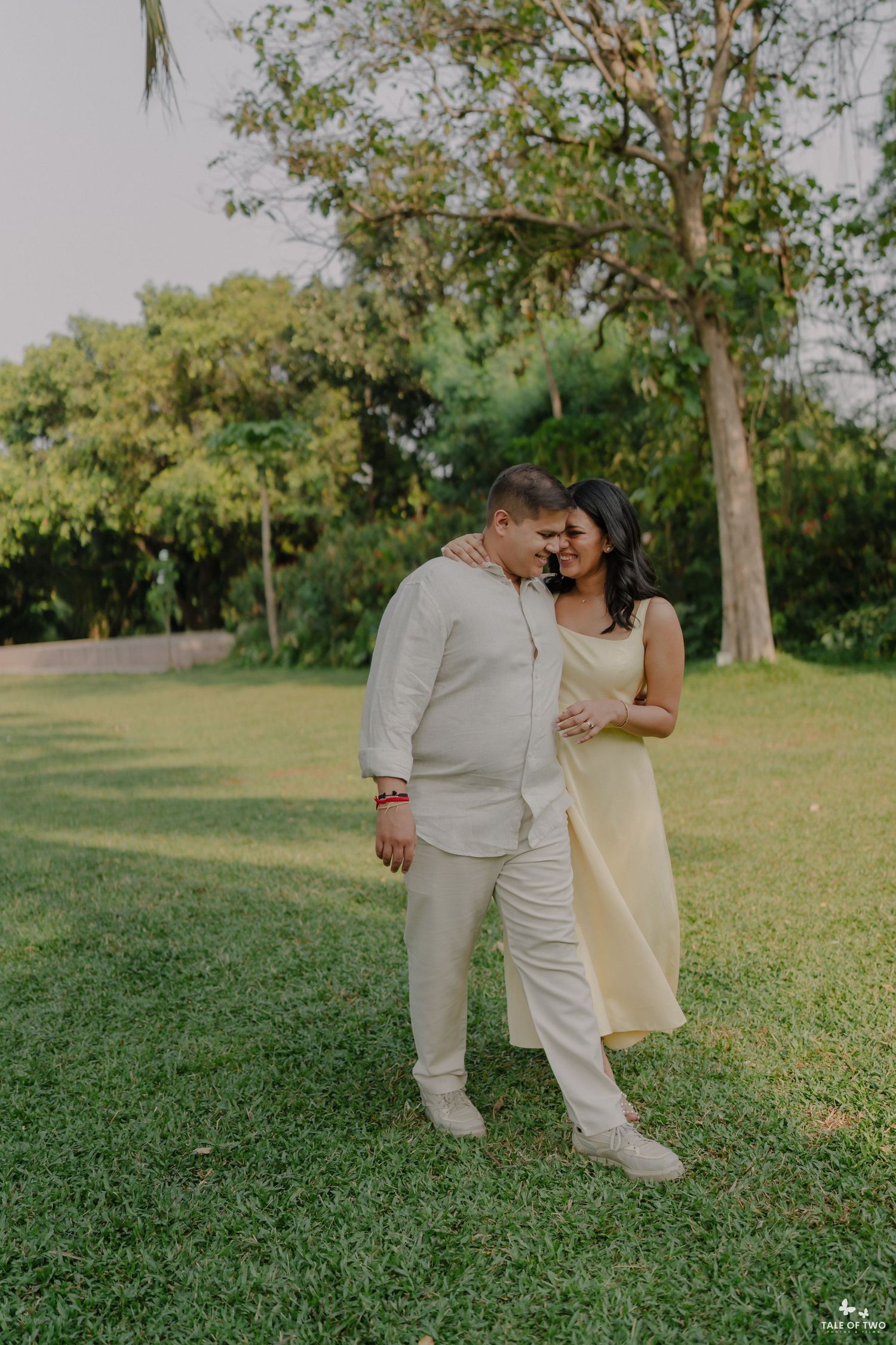 A happy couple in coordinated beige outfits walk together arm in arm on a green lawn with trees.