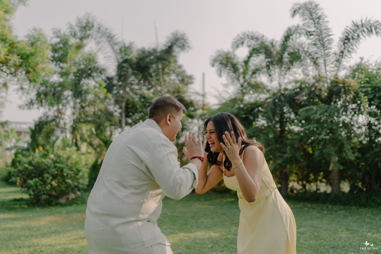 A couple shares a joyful proposal moment outdoors, surrounded by lush green trees and palm trees in a garden setting.