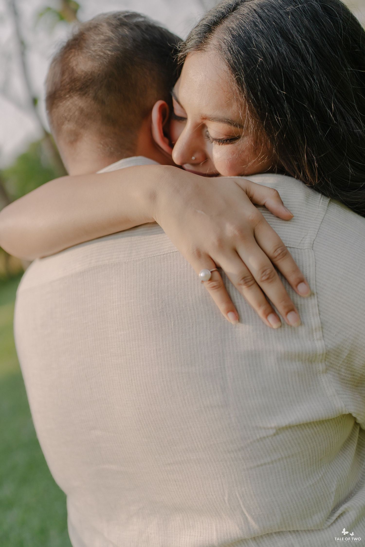 A couple shares a tender embrace outdoors, with the woman displaying an engagement ring on her hand.