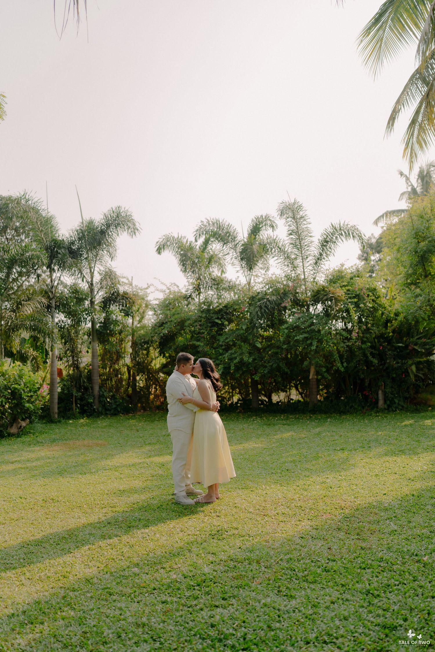 Couple embracing romantically in a lush green garden surrounded by tall palm trees during golden hour.