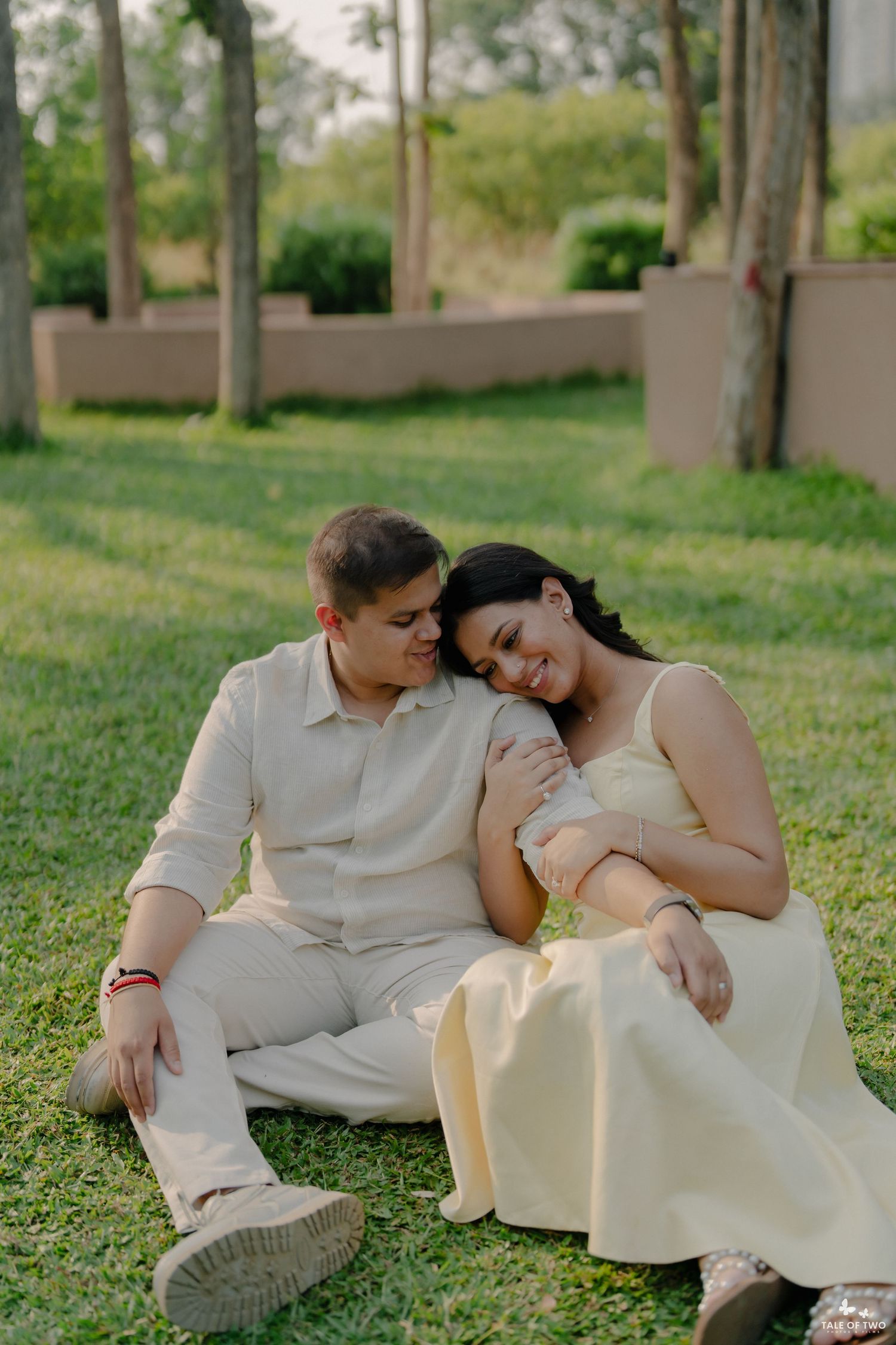 Couple sitting on green grass in a park, woman resting head on man's shoulder, both wearing cream outfits.