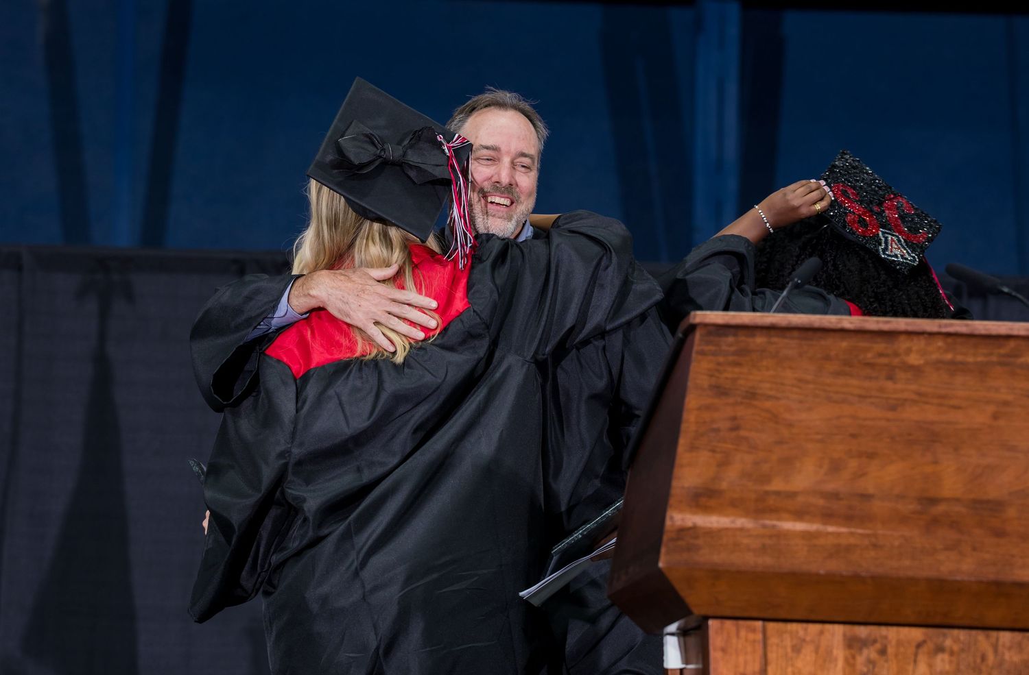 Two graduates in black robes share a warm hug on stage at a graduation ceremony near a wooden podium.