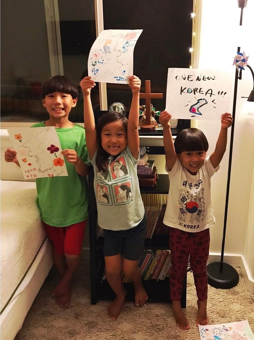 Three young children proudly holding up colorful drawings and signs in a cozy living room at night.