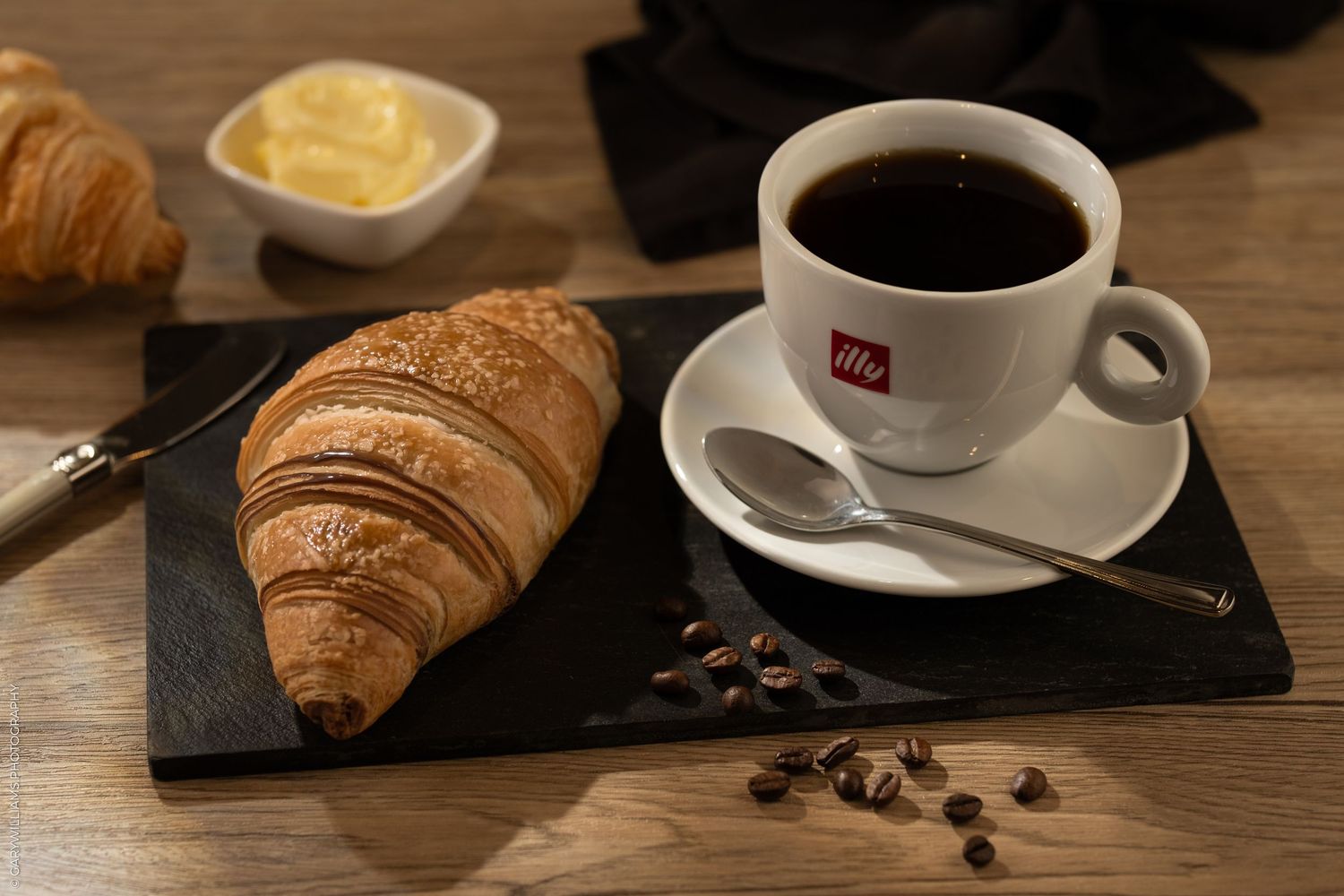 A flaky croissant on a slate board beside an illy coffee cup with scattered beans and butter on a wooden table.