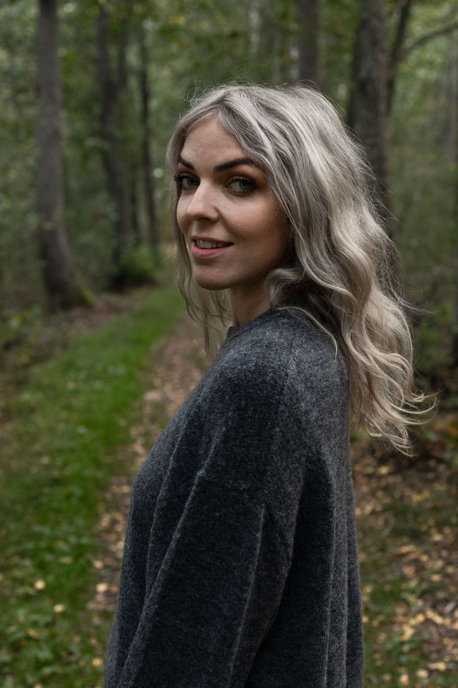 Kvinna i grå tröja poserar i skog med höstlöv och träd i bakgrunden
