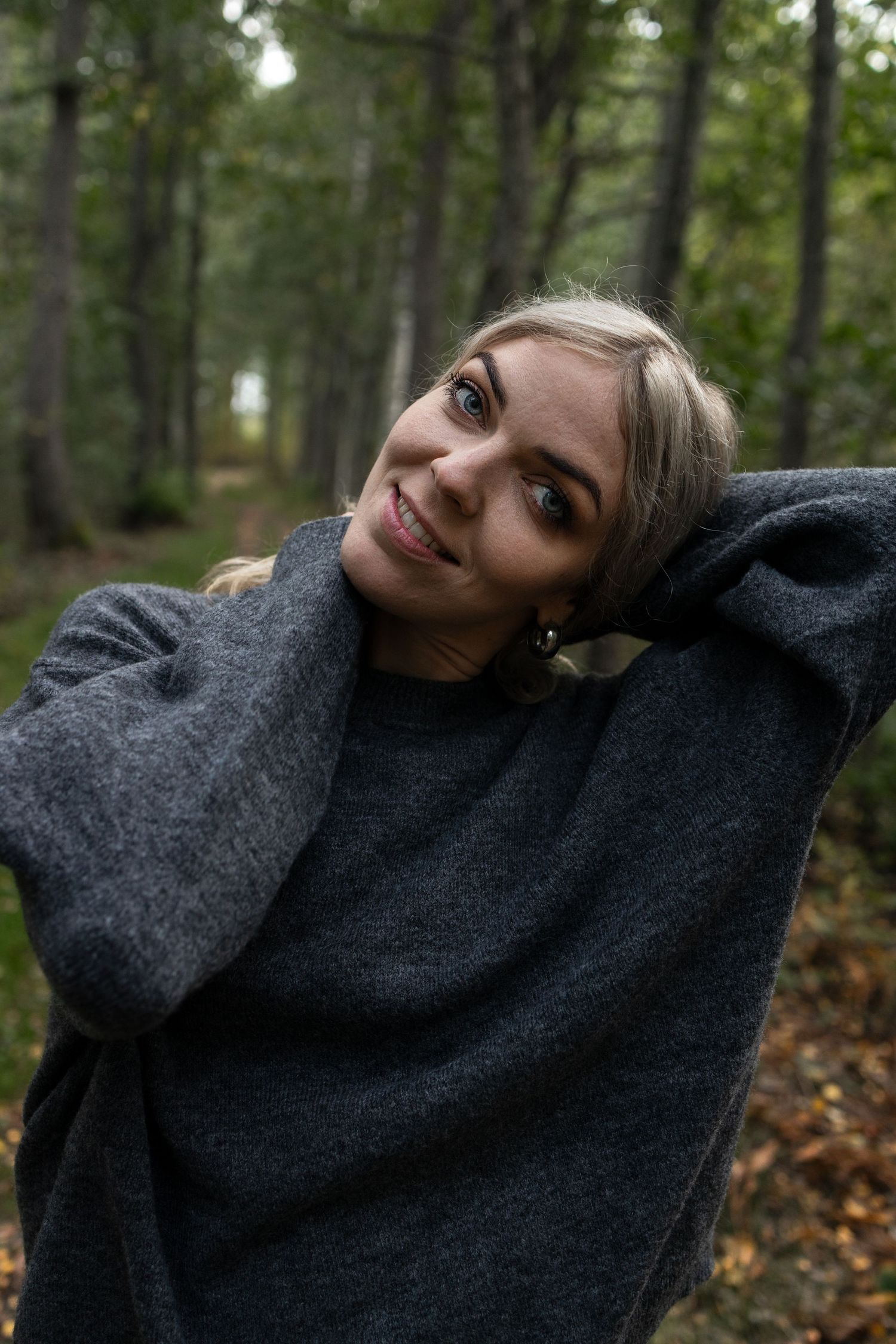 Kvinna i grå tröja ler i höstlig skog med träd och löv i bakgrunden