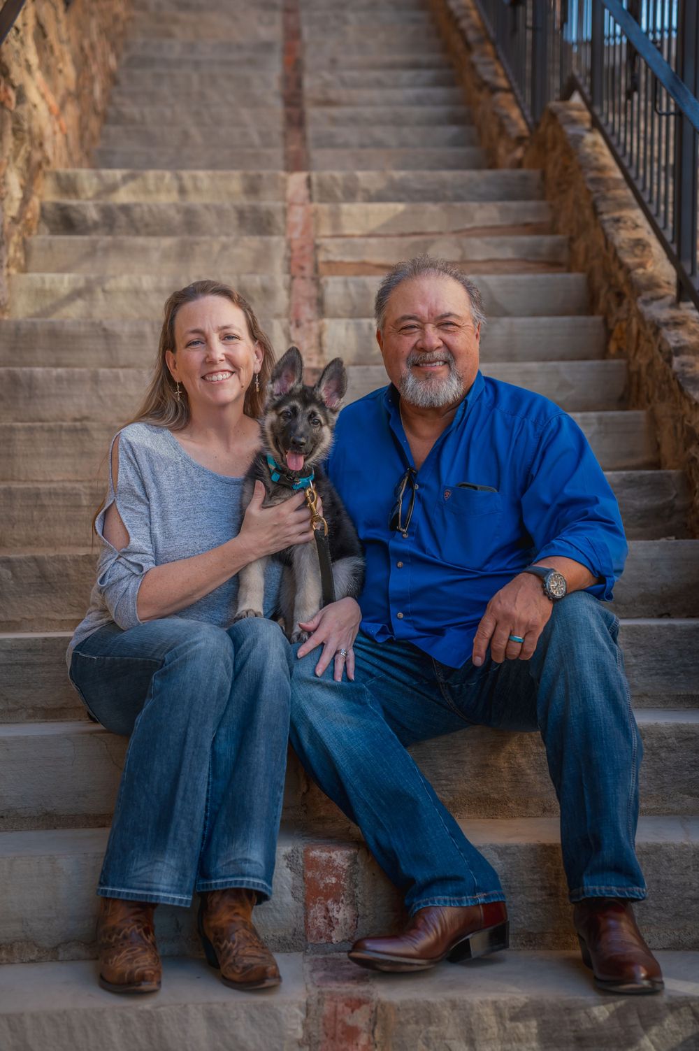 A couple sitting on stone steps with their German Shepherd dog wearing a blue collar.