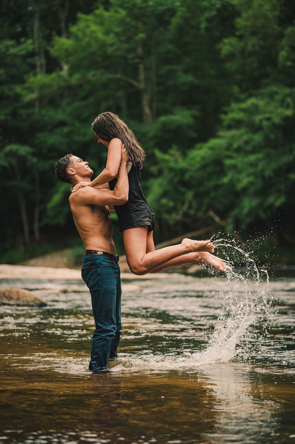 A romantic couple shares an intimate moment in a river as water splashes around them in a forest setting.