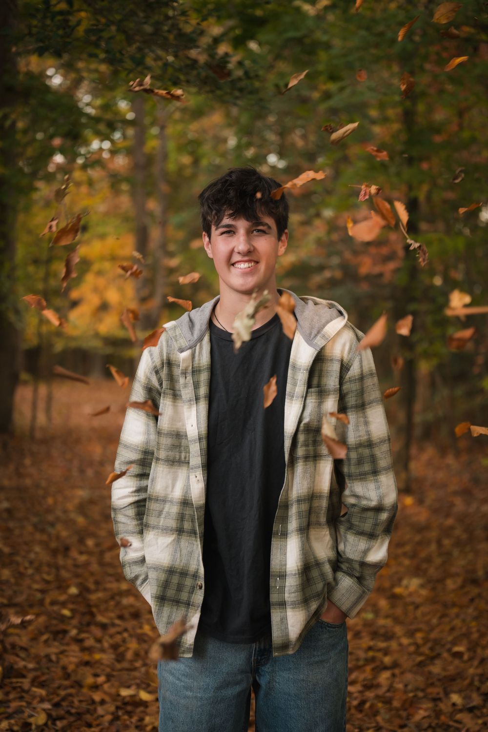 Person wearing plaid flannel jacket and baseball cap poses in autumn forest setting with falling leaves.