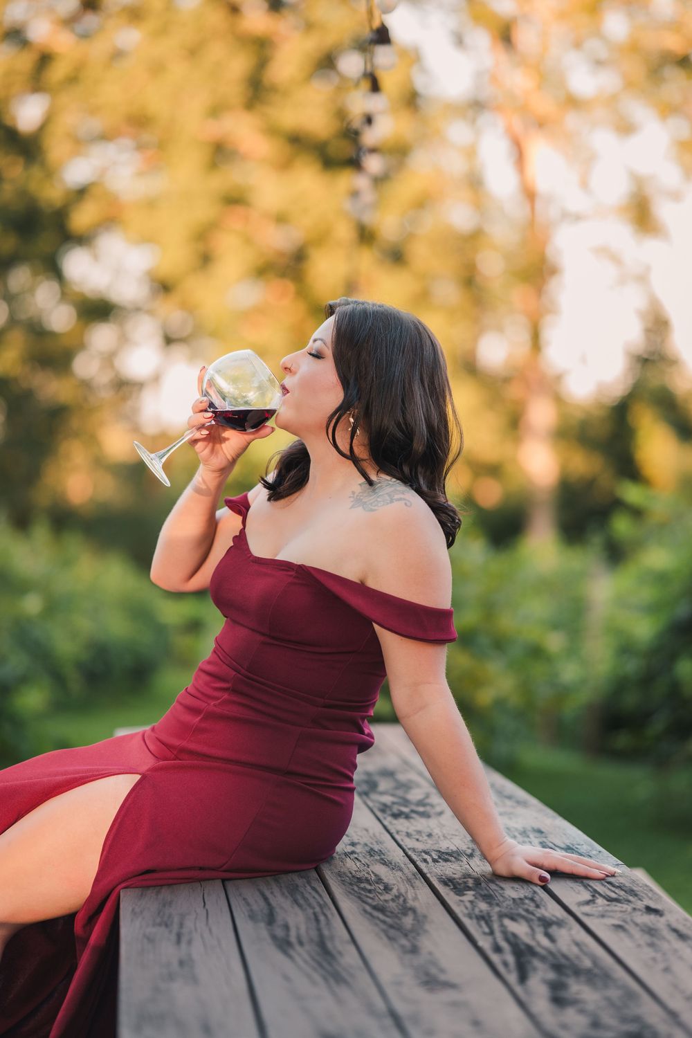 Someone in a burgundy off-shoulder dress sits on a wooden surface sipping wine against an autumn backdrop.