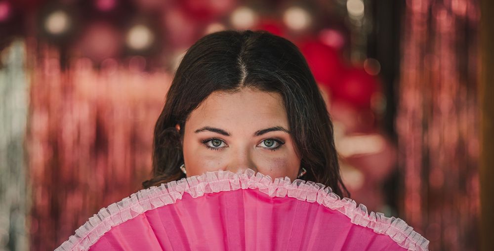 A bright pink feathered fan is held against a red and black illuminated background with bokeh effects.
