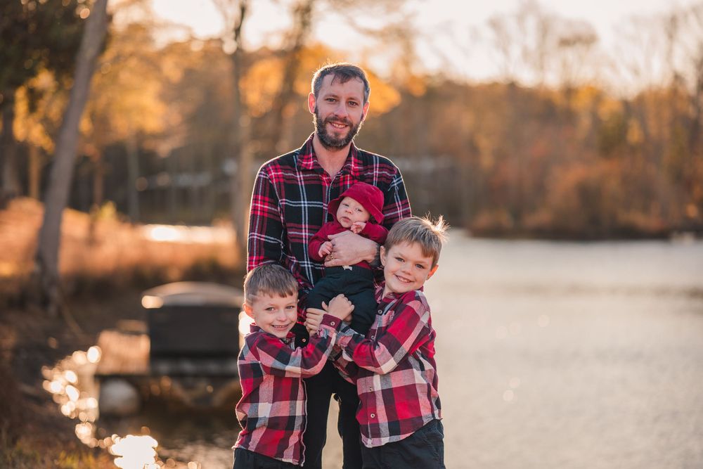 Family wearing matching red plaid outfits poses together during golden hour autumn photoshoot by a lake.