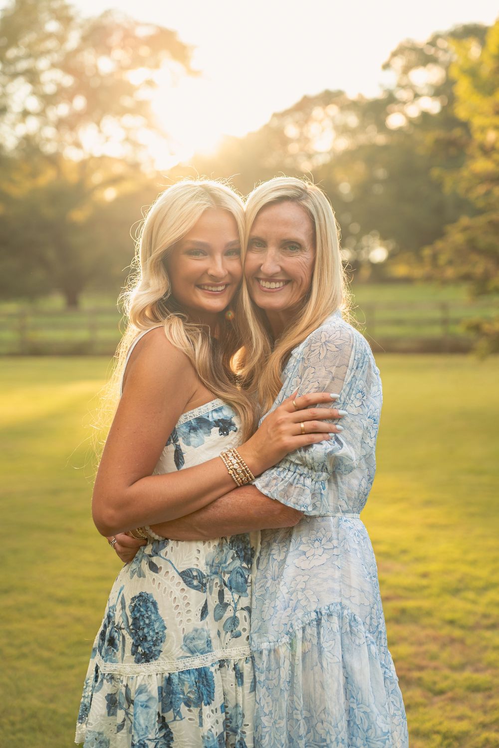 Two people in floral dresses embrace and laugh together in a sunlit field during golden hour.