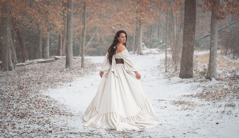 A bride in a flowing white gown poses gracefully in a snowy winter forest setting.