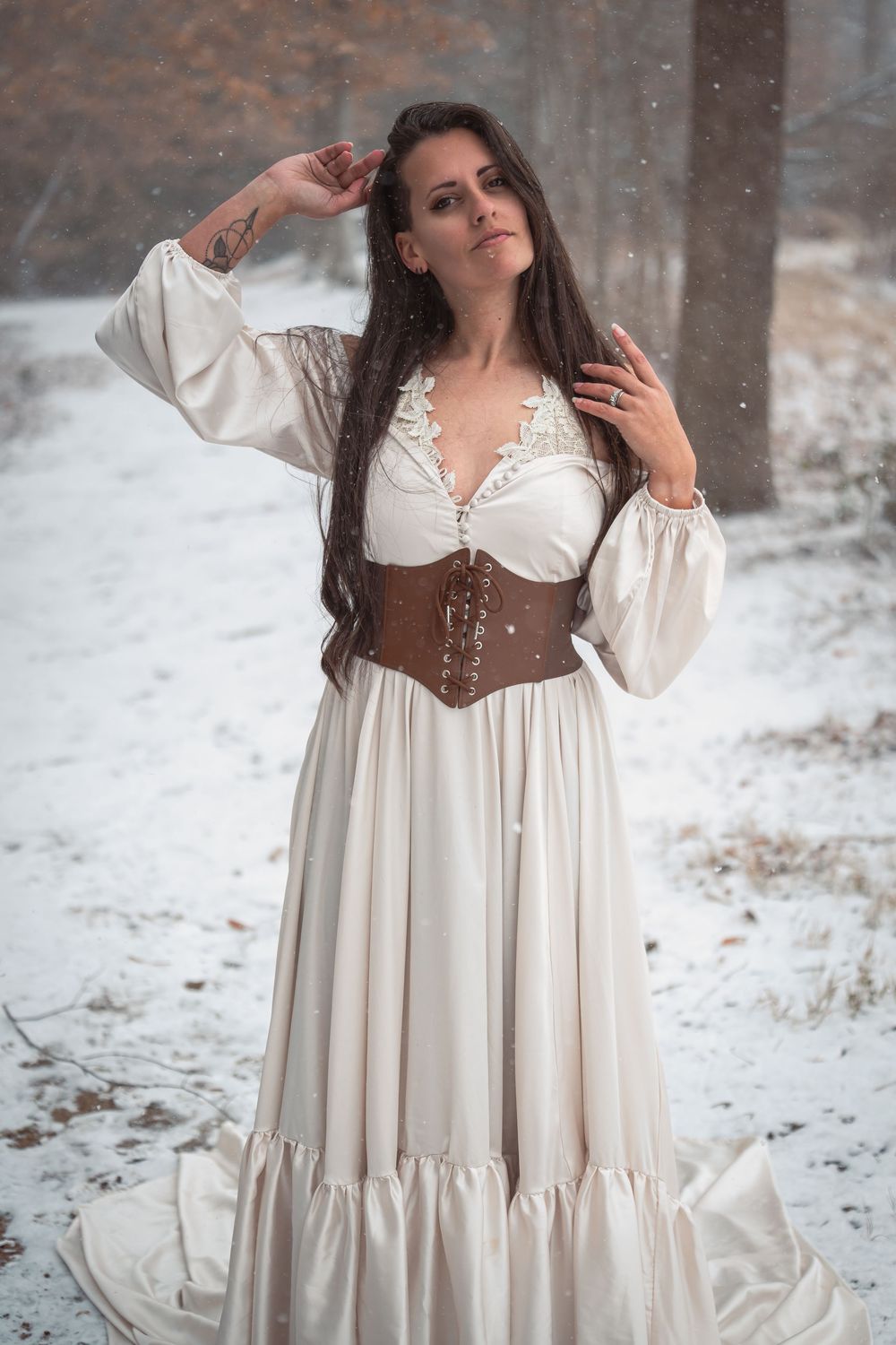 A woman in a medieval-inspired white dress and brown corset poses gracefully in a snowy winter forest setting.
