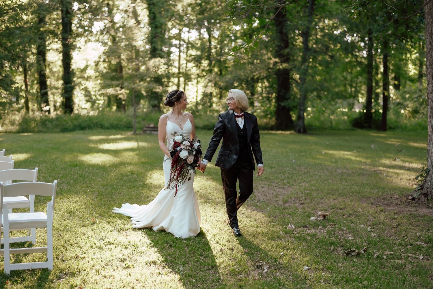 A wedding ceremony scene in a sunlit wooded area with white chairs.