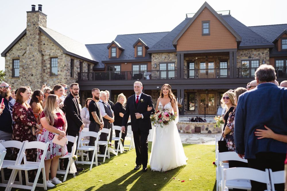A sunny outdoor wedding ceremony in front of a large stone and wood mansion.
