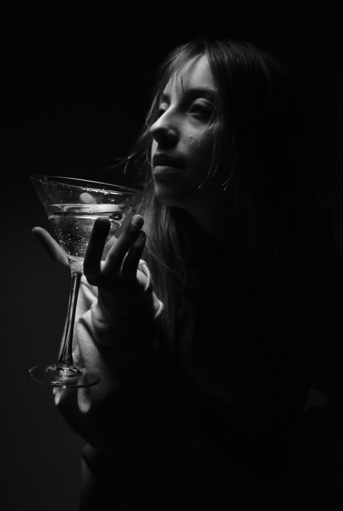 Dramatic black and white portrait of someone holding a martini glass in low light.