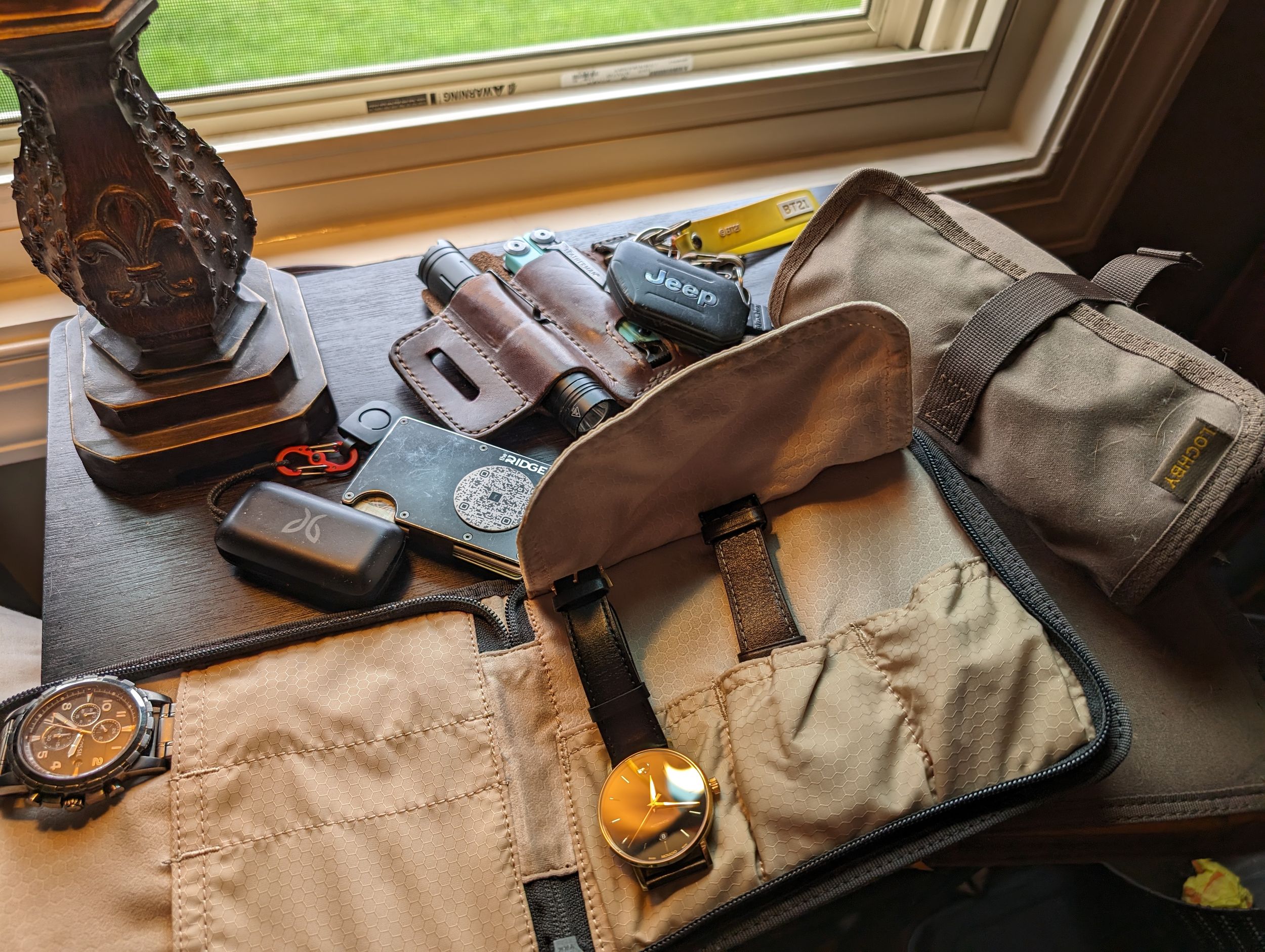 Travel gear and accessories including bags, watch, and electronics laid out on wooden surface near window.
