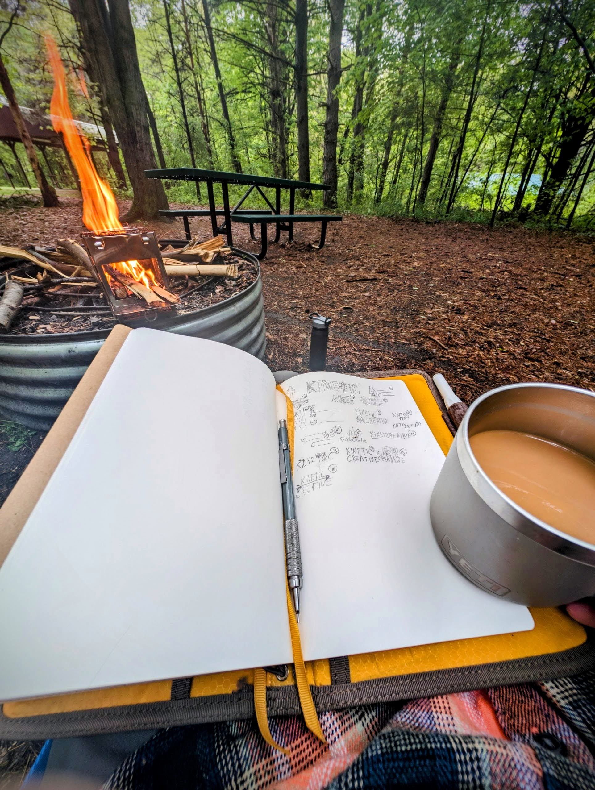 Open notebook and coffee cup next to campfire with picnic table and forest in background.