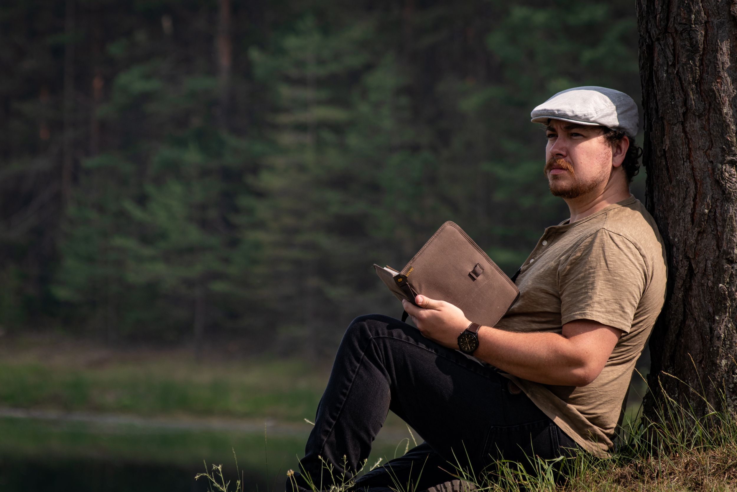 Outdoor scene of someone reading a notebook while sitting against a tree near a lake.