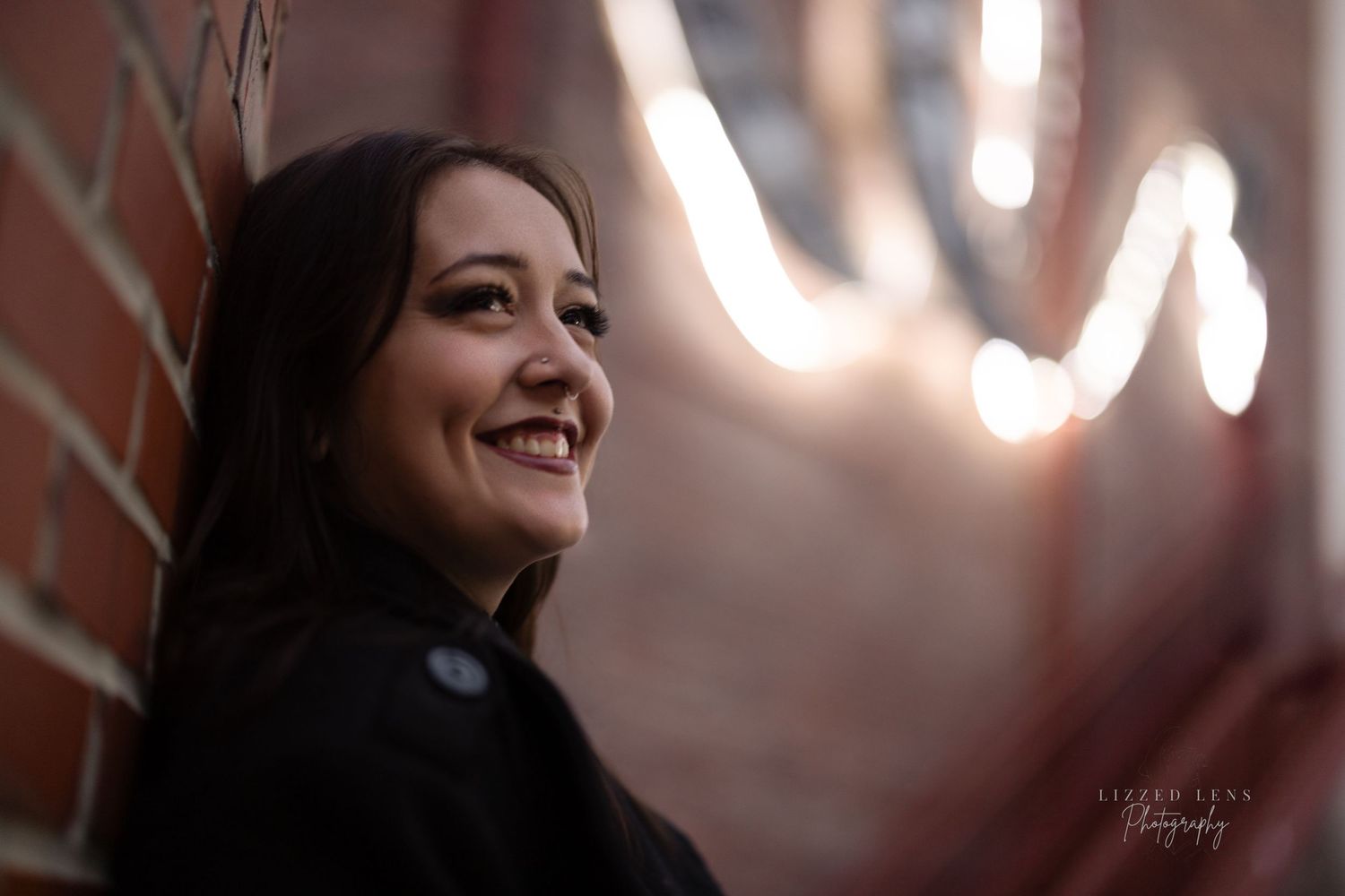 Young woman with dark hair smiling against brick wall with bokeh light background at night.