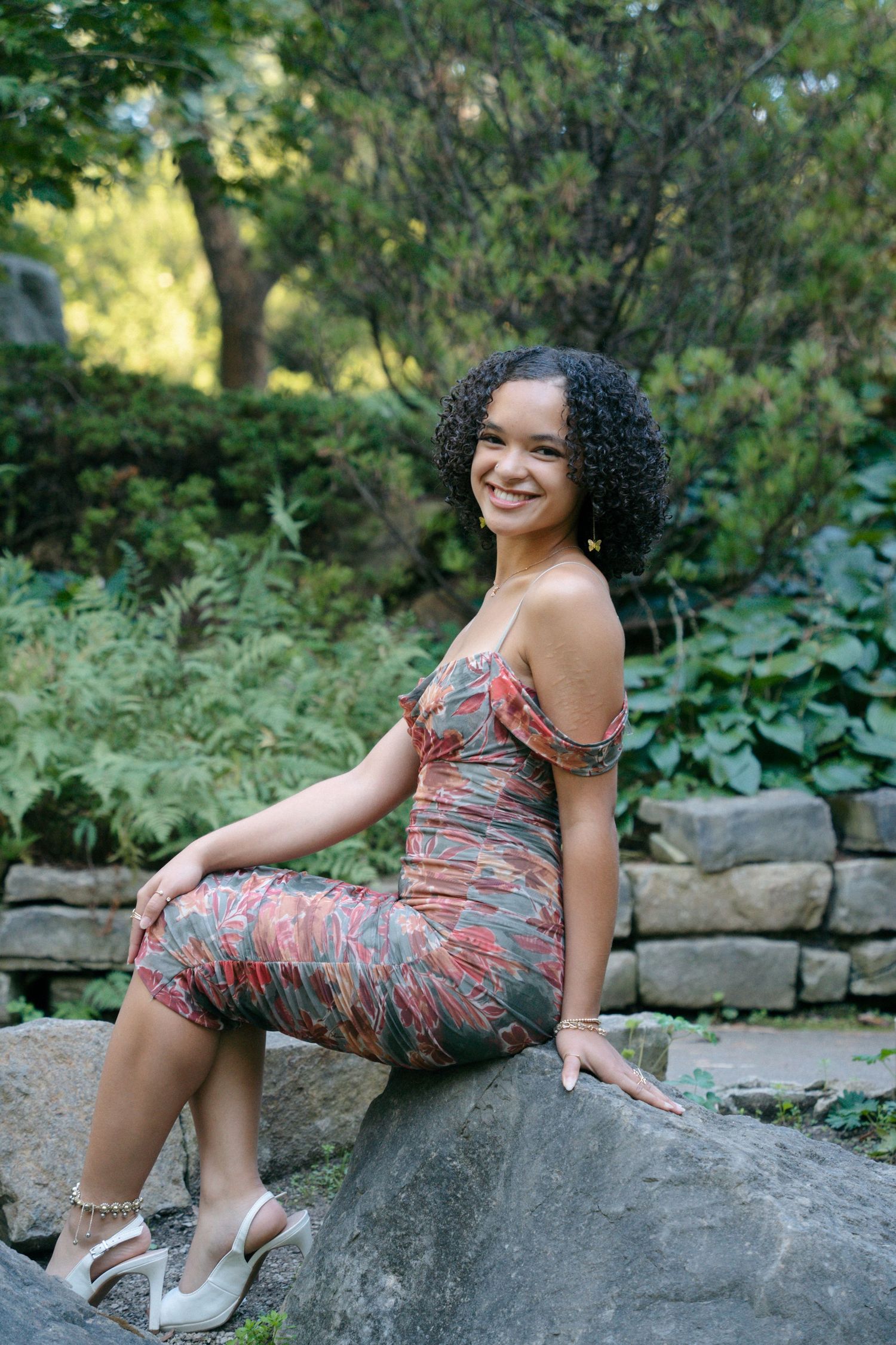 Smiling young woman in floral dress sits on rocks in a lush green garden setting.