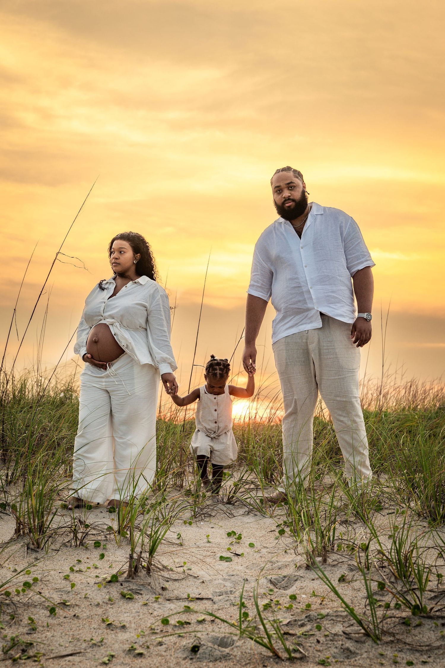 Family wearing white outfits poses at sunset on sand dunes with beach grass.