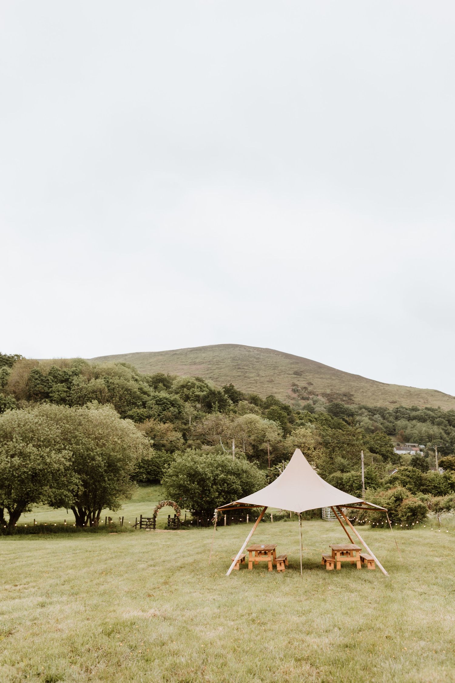 A beige stretch tent set up in a green field with wooden furniture beneath, surrounded by hills and trees in nature.