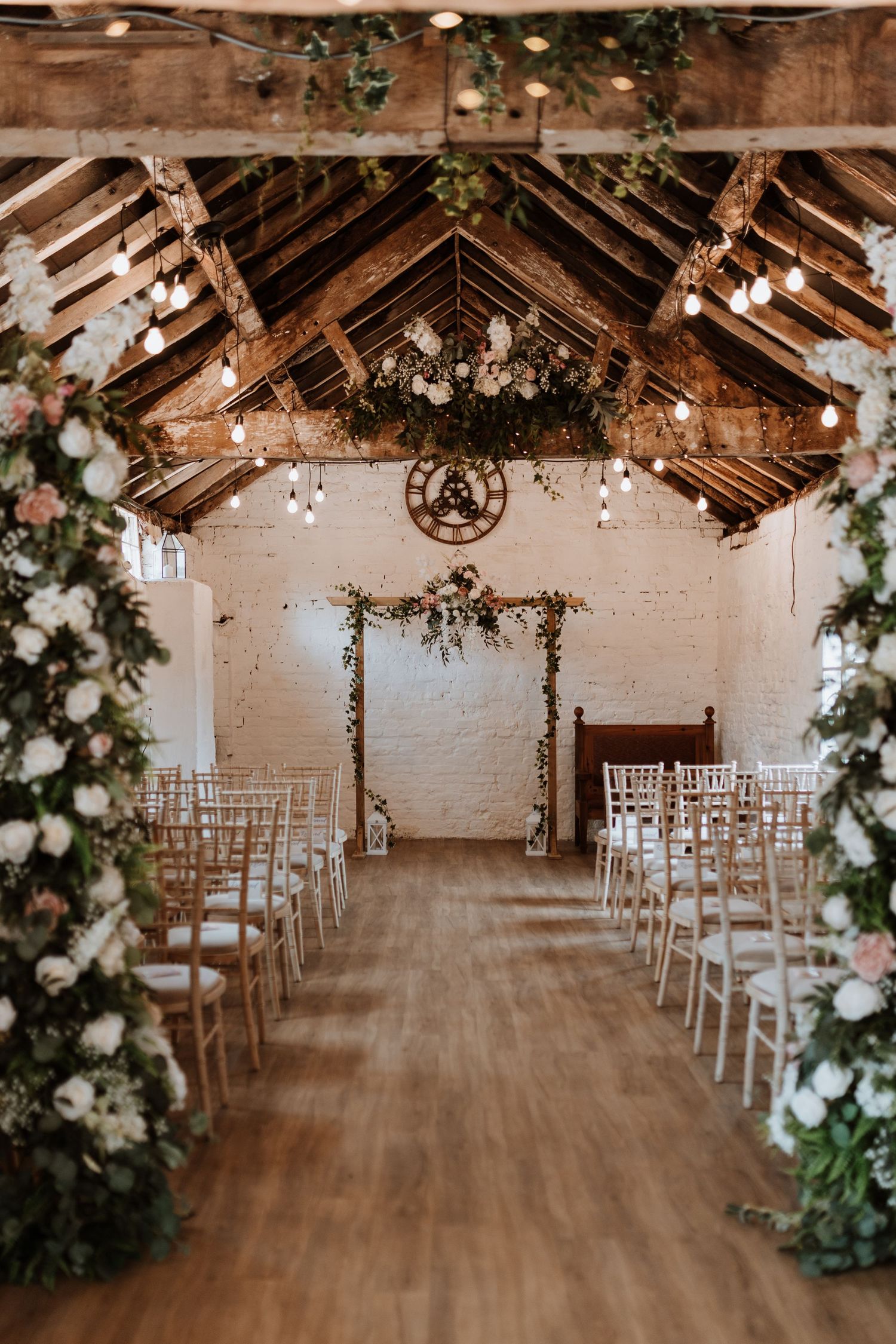 Rustic barn wedding ceremony setup with white floral arch, wooden beams, hanging lights and white chiavari chairs.