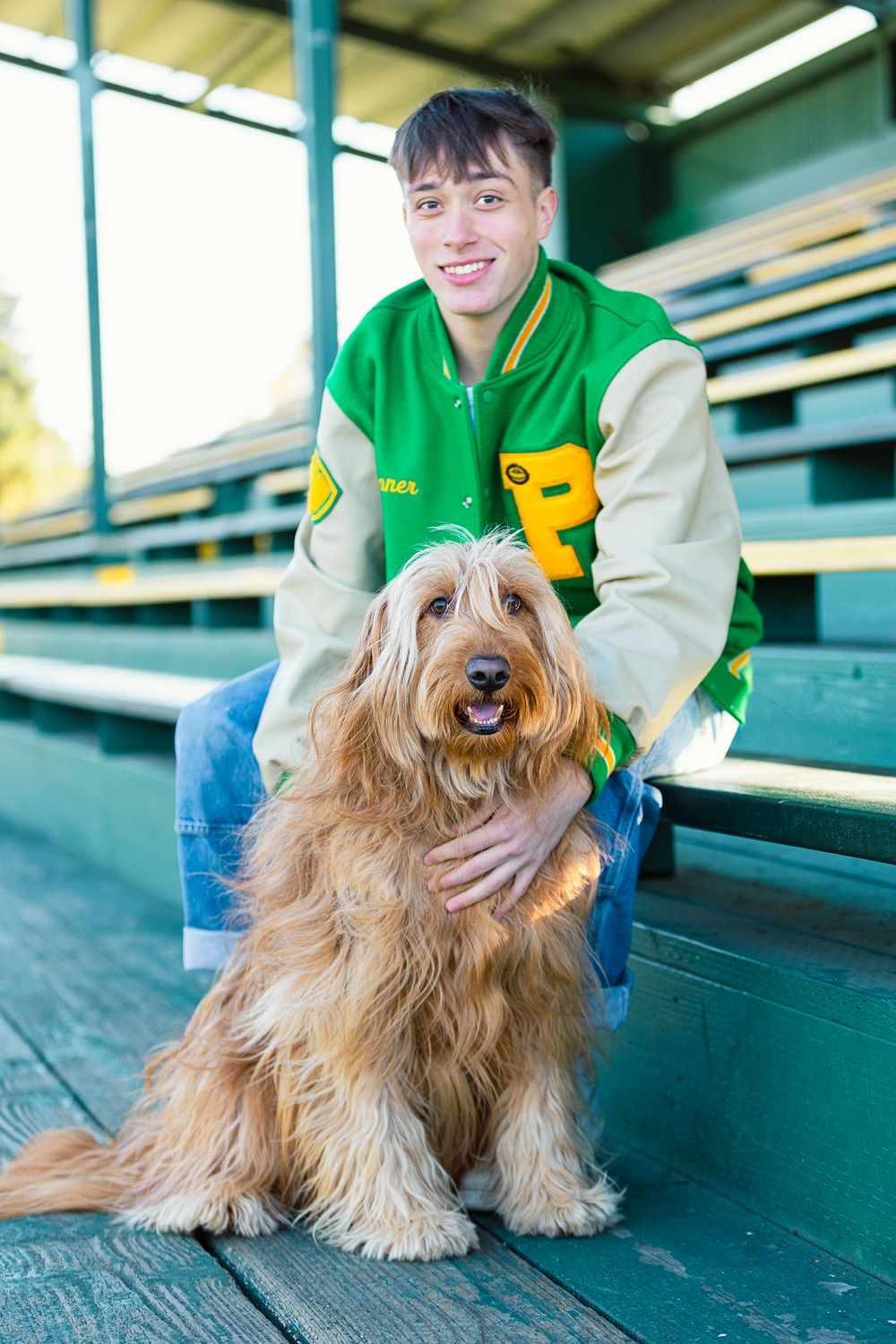 A fluffy brown Tibetan Terrier dog sits on bleachers with someone wearing a green and white varsity jacket.