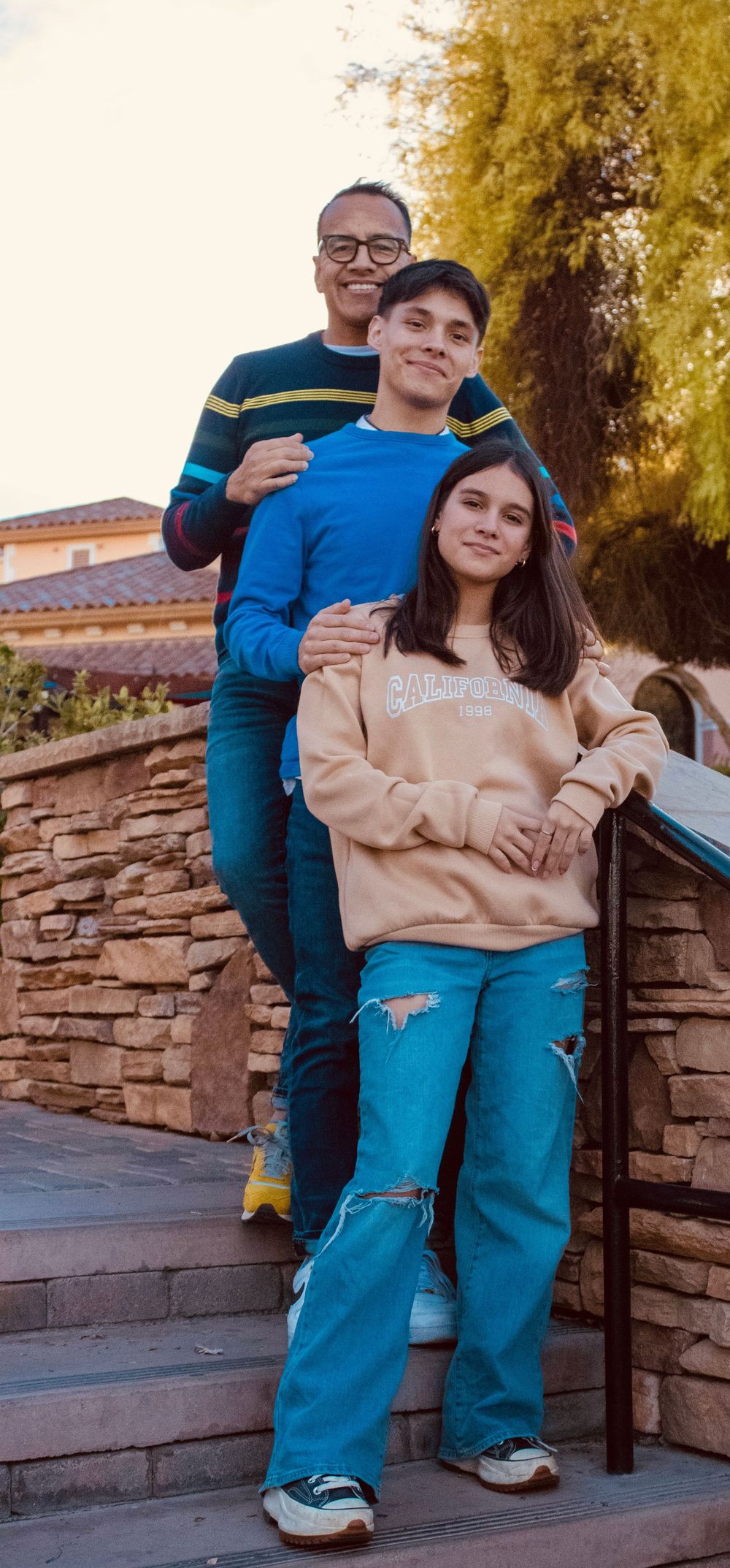A father and two teenagers pose on stone steps outside a building surrounded by autumn trees.
