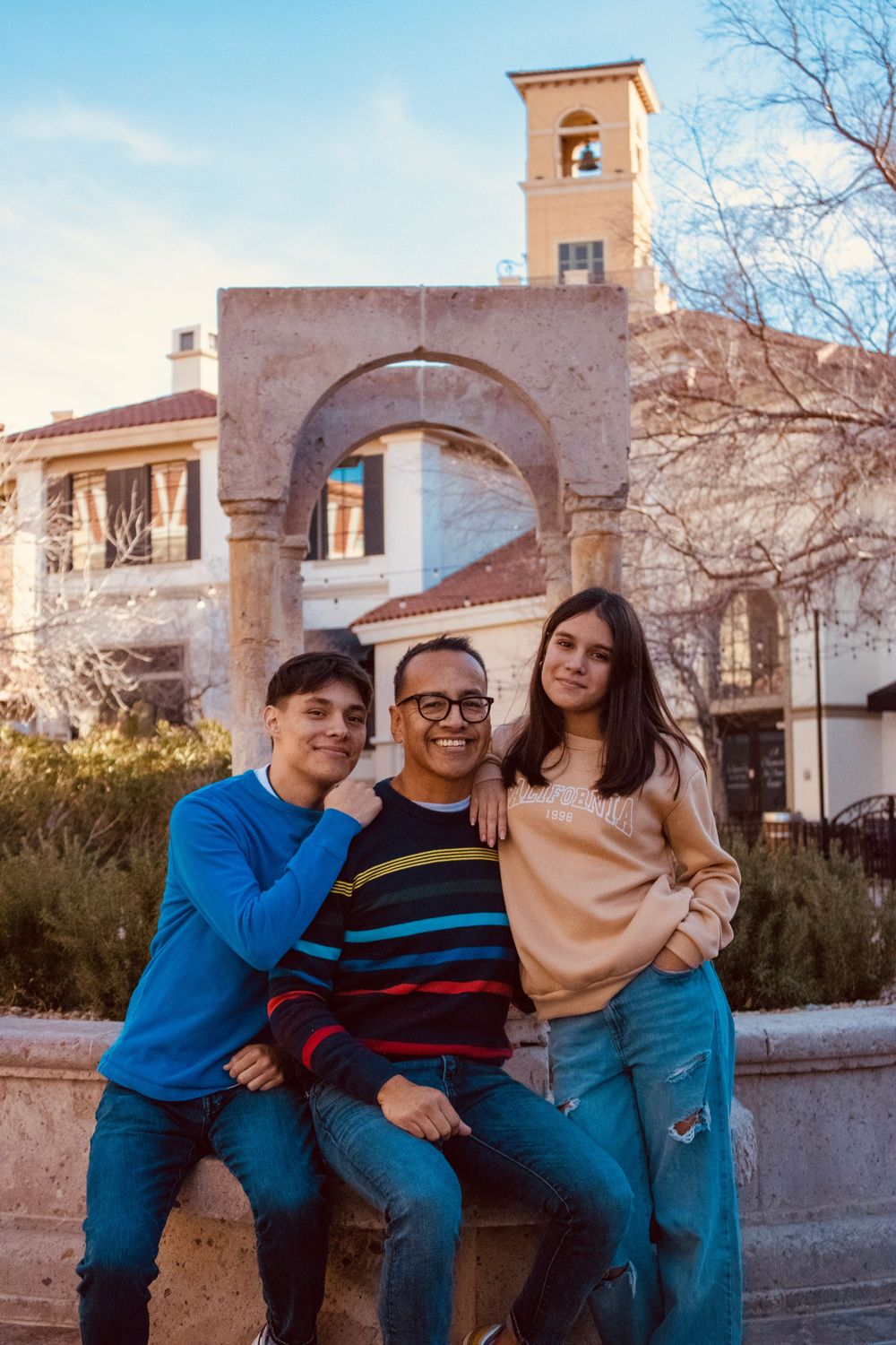 Father posing with teenage son and daughter in front of stone arch at outdoor shopping plaza at sunset.
