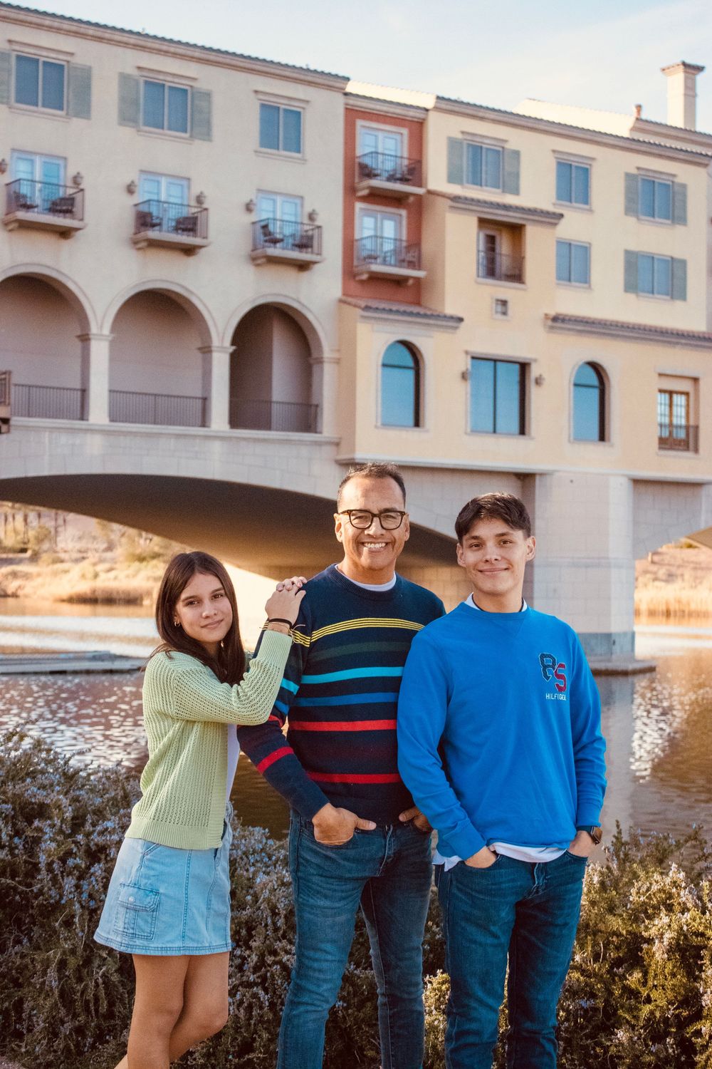 Father smiling with teenage daughter and son standing by waterway at outdoor shopping center at sunset.